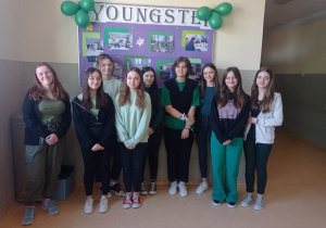 1.Na zdjęciu widać grupę nastoletnich, uśmiechniętych dziewcząt, ubranych w zielone barwy na korytarzu szkolnym.