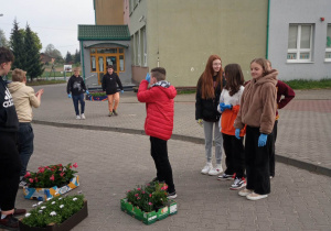 10.Zdjęcie ukazuje grupę nastoletnich dzieci, stojących na dziedzińcu szkoły. Młodzież przygotowuje się do sadzenia roślin.