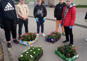 14.Zdjęcie ukazuje grupę nastoletnich dzieci, stojących na dziedzińcu szkoły. Młodzież przygotowuje się do sadzenia roślin.