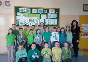 16.Na zdjęciu widzimy grupkę młodszych dzieci ubranych w zielone barwy na korytarzu szkolnym pod ekologiczną gazetką wraz z dwiema nauczycielkami.