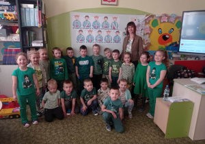 18.Zdjęcie ukazuje grupę przedszkolaków wraz z opiekunem. Wszyscy są ubrani w zielone barwy i znajdują się w Sali przedszkolnej.