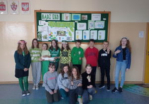7.Na zdjęciu widzimy grupkę młodszych dzieci ubranych w zielone barwy na korytarzu szkolnym pod ekologiczną gazetką.