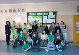 8.Na zdjęciu widzimy grupkę młodszych dzieci ubranych w zielone barwy na korytarzu szkolnym pod ekologiczną gazetką wraz z dwiema nauczycielkami.