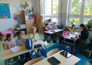 Uczniowie klasy 3 siedzą skupieni w ławkach. Dziewczynki z klasy 6 pokazują sposób mnożenie dwóch liczb na palcach.
