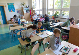 Uczniowie siedzą w ławkach. Każdy uczeń ma tablet i gra w turnieju z tabliczki mnożenia. Przy każdym stoliku jest uczennica klasy 6.