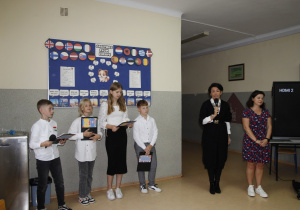 Na zdjęciu widnieje dyrektor szkoły, nauczyciel języka angielskiego oraz grupka dzieci z mikrofonami na tle gazetki.
