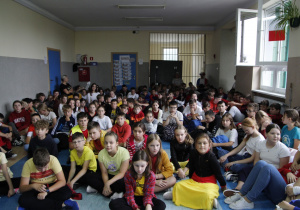 zdjęcie ukazuje grupę siedzących na korytarzu szkolnym dzieci wraz z nauczycielami