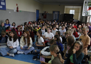 zdjęcie ukazuje grupę siedzących na korytarzu szkolnym dzieci wraz z nauczycielem, które rozwiązują quiz językowy