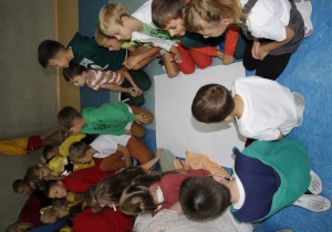 zdjęcie ukazuje grupę siedzących na korytarzu szkolnym dzieci wraz z nauczycielem, które pracują nad brystolem