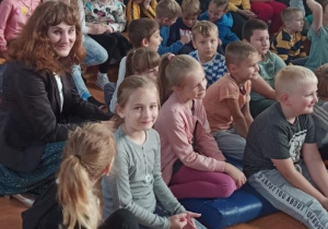 Uczniowie klas I-IV i klasy VII uczestniczą w spotkaniu autorskim z Łukaszem Wierzbickim. Na pierwszym planie wraz z uczniami siedzi Monika Fau. Uśmiecha się lekko.