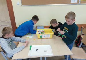 Dwie złączone ławki szkolne. Na nich pudełko z klockami Lego Spike i tablet. Przy ławkach czterech chłopców buduje z klocków młyn.