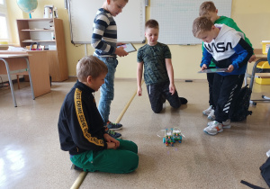 Sala lekcyjna. Po podłodze jadą dwa roboty. Pięciu chłopców obserwuje poruszanie się robotów.