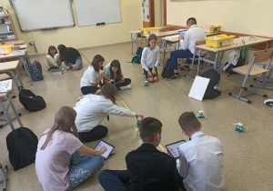 Sala lekcyjna. Uczniowie grupami siedzą na podłodze. W rękach trzymają tablety na których programują poruszanie się robotów. Zaprogramowane roboty poruszają się po podłodze.