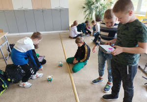 Sala lekcyjna. Uczniowie grupami siedzą na podłodze i obserwują jeżdżące roboty. Dwóch chłopców stoi, jeden z nich w ręku trzyma tablet na którym programuje poruszanie się robota.
