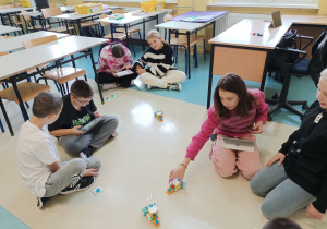 Sala lekcyjna. Na podłodze dwójkami siedzą uczniowie. Na tabletach kodują poruszanie się robotów.