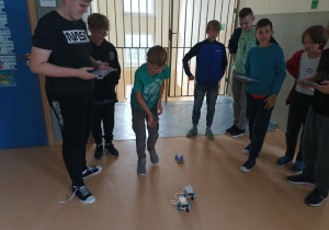 Korytarz szkolny. Grupa chłopców obserwuje wyścig robotów. Uczniowie w rękach trzymają tablety na których programują kierunek i prędkość poruszania się robotów. Roboty zostały zbudowane z klocków lego.