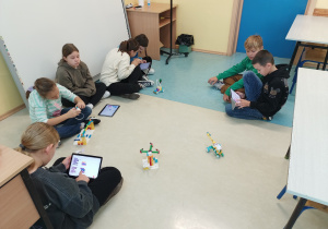 Sala lekcyjna. Na podłodze siedzi grupa uczniów, przed nimi roboty. Uczniowie w rękach trzymają tablety i programują poruszanie się robotów.