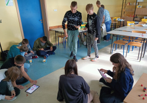 Sala lekcyjna. Siedmiu uczniów siedzi na podłodze, czterech stoi. Uczniowie trzymają tablety na których programują swoje roboty. Na twarzach widać zadowolenie z osiągniętego celu.