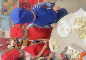 Dwie dziewczynki w czerwonym stroju kucharskim oraz jeden chłopiec kroją banany.