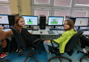 Dwie uczennice w klasopracowni informatycznej siedząc z uśmiechem na twarzy oczekują na ocenę swoich prac. W tle na ekranach monitorów widoczne są efekty ich prac. Dziewczynki odkodowały krzesła na polanie.
