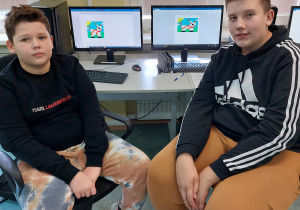 Przy swoich stanowiskach w klasopracowni informatycznej siedzi dwóch uczniów. W tle na ekranach monitorów widoczne są wykonane przez nich prace. Uczniowie odkodowali obrazek przedstawiający psa.