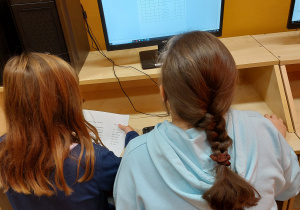 Dwie uczennice siedząc przy stanowisku komputerowym wykonuje pracę polegającą na kodowaniu obrazka. W tle na ekranach monitorów widoczne są początkowe efekty pracy.