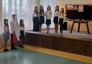 Scena, na niej pięć uczennic. Za nimi tablica na której widnieje biało-czerwona flaga oraz godło Polski. Przed sceną, z lewej strony stoi poczet flagowy.