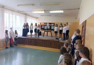 Scena, na niej uczniowie śpiewają utwór pt. „Biały krzyż”. Przed sceną uczniowie klasy 1 i 2 wsłuchani w słowa pieśni. Po lewej stronie poczet flagowy.