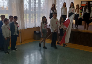 Uczeń z pochyloną flagą, obok niego asysta, dwie uczennice wyprowadzają poczet flagowy.