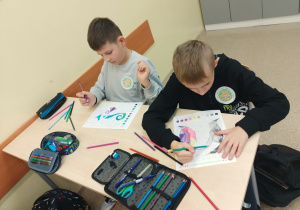 Ławka szkolna, na niej piórniki i kredki. Dwóch chłopców siedzi i koloruje obrazek według kolorów zakodowanych działaniami z tabliczki mnożenia.
