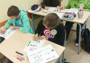 Sala lekcyjna, przy stolikach siedzą uczniowie i wykonują działania z tabliczki mnożenia, by prawidłowo pokolorować rysunki.