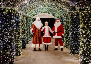 Mikołaj oraz jego pomocnicy w czerwonych ubraniach witają stoją w tunelu światełek.