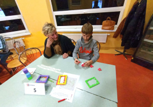 Złączone stoliki szkolne, przy nich mama z synem. Mama obserwuje, jak syn próbuje ułożyć sześcian z czerwonych puzzli Happy Cube.