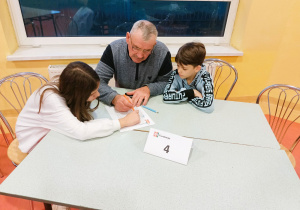Zsunięte stoliki szkolne, przy nich dziadek z wnuczką i wnukiem wśród zapisanych na kartce liter szukają i wykreślają wszystkie wyrazy związane z matematyką.