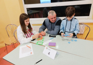 Zsunięte stoliki szkolne, przy nich dziadek z wnuczką i wnukiem składają sześciany z kolorowych puzzli Happy Cube.