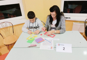 Zsunięte stoliki szkolne, przy nich mama z synem składają sześciany z kolorowych puzzli Happy Cube.