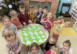Grupa dzieci przedszkolnych pozuje do zdjęcia z wykonanymi kartkami świątecznymi.