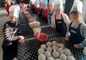 Dziewczynki w czapkach kucharskich i fartuszkach przy stole, na którym znajdują się produkty do przygotowania pizzy.