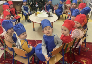 grupa dzieci przedszkolny ubranych w zestawy kucharskie w kolorze czerwonym i niebieskim siedzą na krzesełkach.