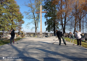 na zdjęciu widać czworo sprzątających dzieci oraz drewniany krzyż na wybrukowanym placu