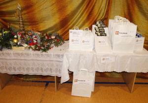 Stolik nakryty białym obrusem. Na stoliku torebki z nagrodami dla uczestników projektu. Na torebkach logo programu napis mFundacja.