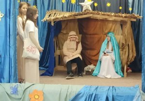 Szopka, przed którą stoi grupa osób. W szopce siedzi chłopiec w przebraniu Józefa i dziewczynka przebrana za Maryję.