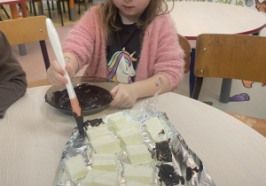 Dziewczynka smaruje pędzelkiem zamoczonym w czekoladzie białe prostokąty ptasiego mleczka.