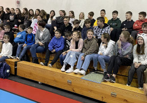 uczniowie siedzą na trybunach na hali sportowej i oglądają występ