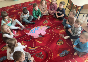 Dzieci siedzą na dywanie a w środku leży dinozaur.