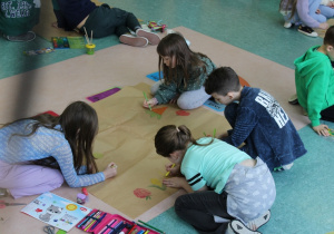 Grupy dzieci malują owoce, warzywa i kwiaty na dużych, rozłożonych na podłodze kartkach. Dzieci siedzą dookoła nich. Rysunki są kolorowe i duże. Uczniowie są zaangażowani w tworzenie prac.