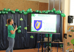 : na zdjęciu widzimy panią w zielonym stroju, w tle zielone balony, jest tam także tablica interaktywna