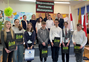 wspólna fotografia wszystkich nagrodzonych uczniów w konkursie pod tytułem Kochajcie Ojczystą Ziemię, nad którymi znajduje się tablica z napisem Wojewódzkie Konkursy Historyczne.