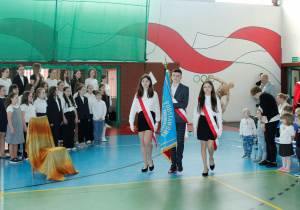 Zdjęcie ukazuje uczniów niosących sztandar szkoły w hali sportowej, w tle widzimy uczniów stojących na baczność.
