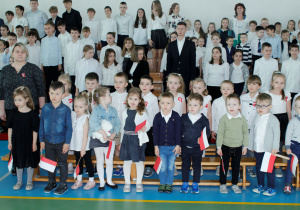 Na zdjęciu widzimy dzieci w wieku przedszkolnym, które stoją w galowych strojach w hali sportowej. Dzieci trzymają w rękach biało- czerwone flagi.
